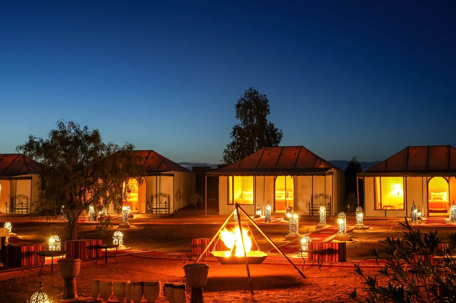 The evening campfire burning in the foreground with the lit tents of the camp behind at dusk