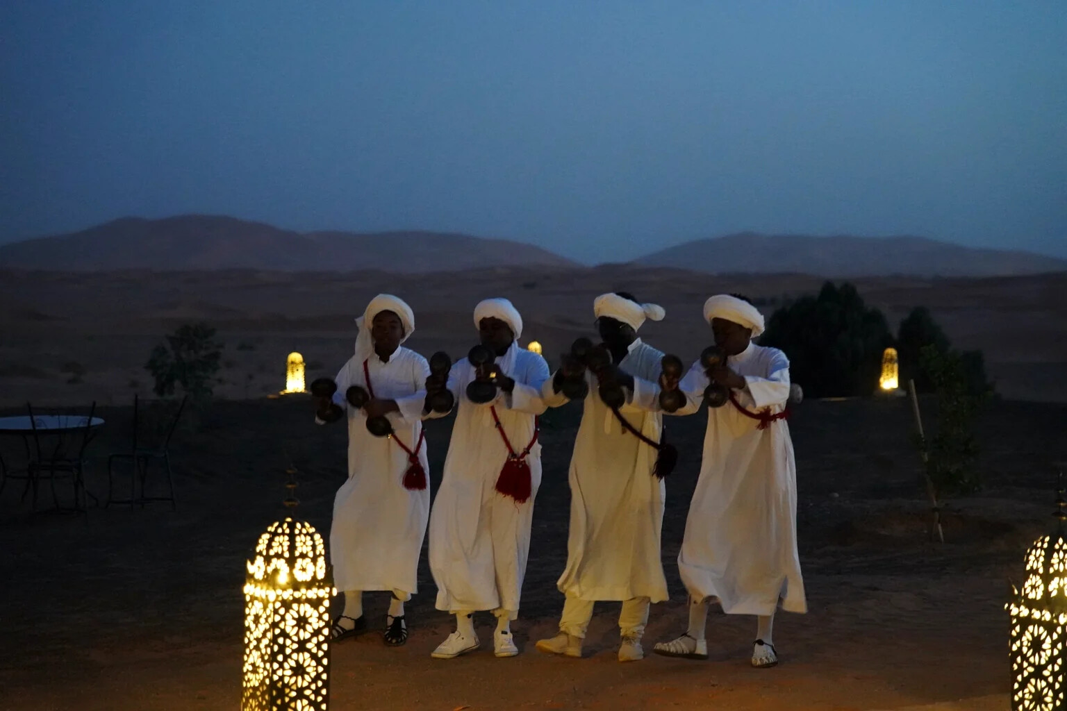 Four Gnawa musicians in white robes playing metal qraqeb beside lanterns at dusk in the desert