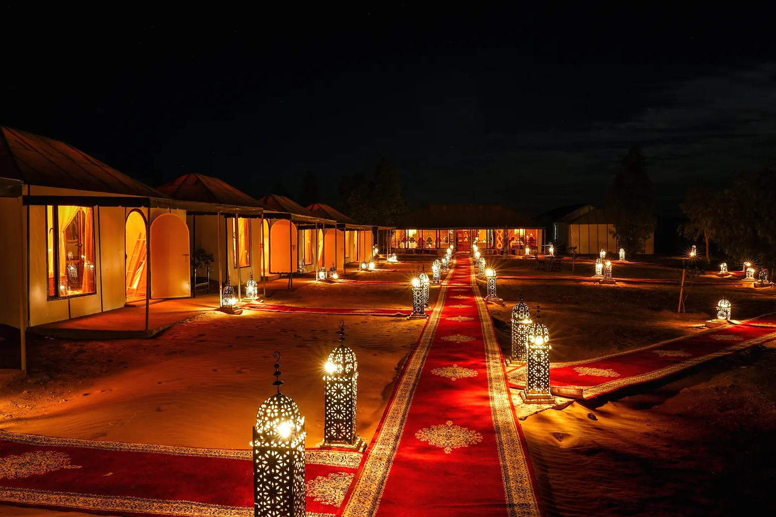 Looking down the lantern-lit red carpet path at night with dark dunes on the horizon
