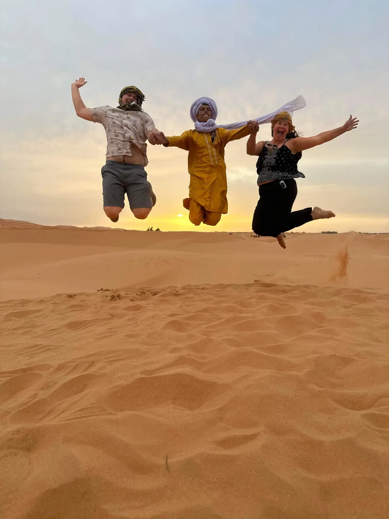 Two guests and their desert host jumping together on a dune at sunset near Merzouga