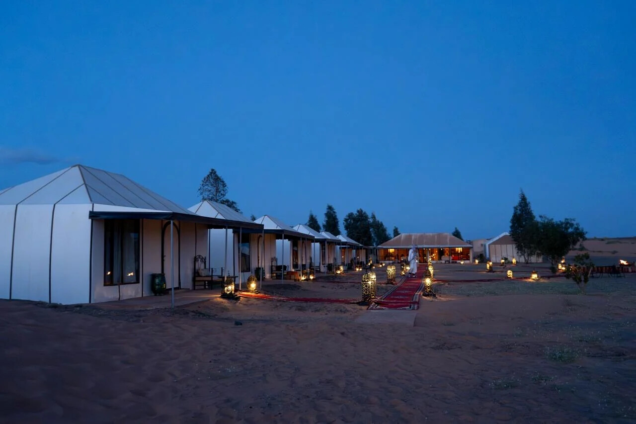 Wide view of the desert camp at dusk with lanterns lighting the red carpet path between the tents