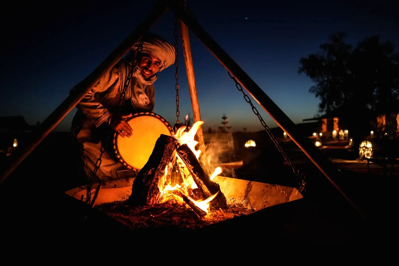 A musician silhouetted against the campfire playing a hand drum under the desert night sky