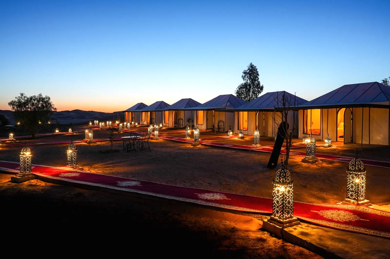 The camp tents and lantern-lit walkways seen across the sand at blue hour