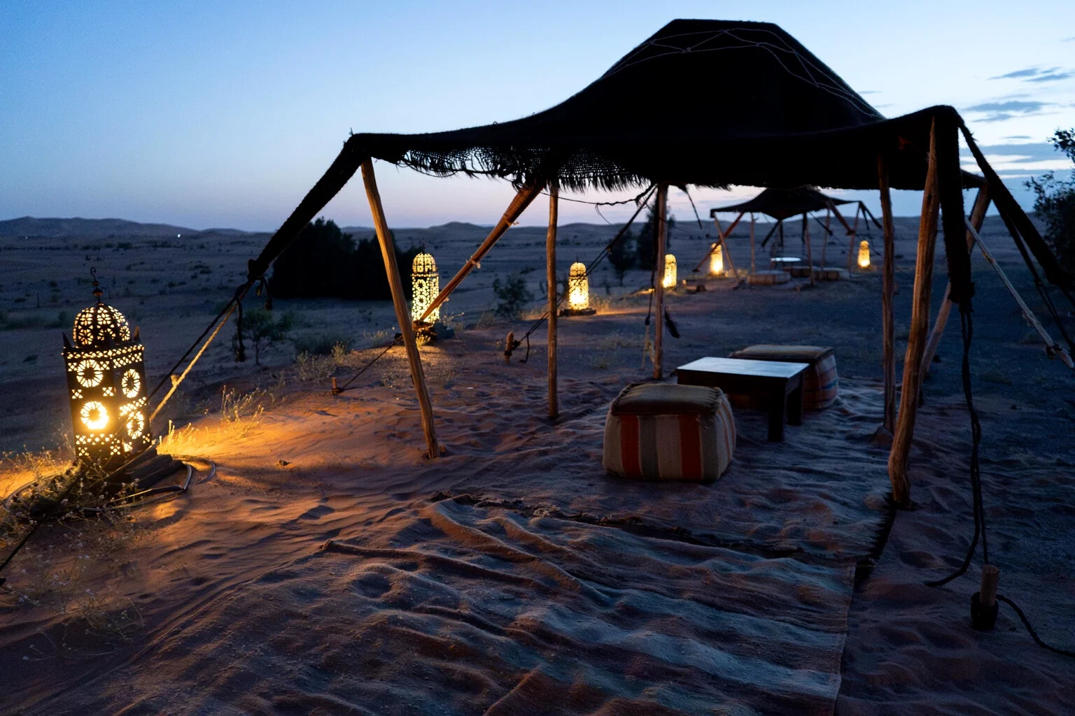 Black Berber canopy over a sand lounge lit by Moroccan lanterns at blue hour near Merzouga