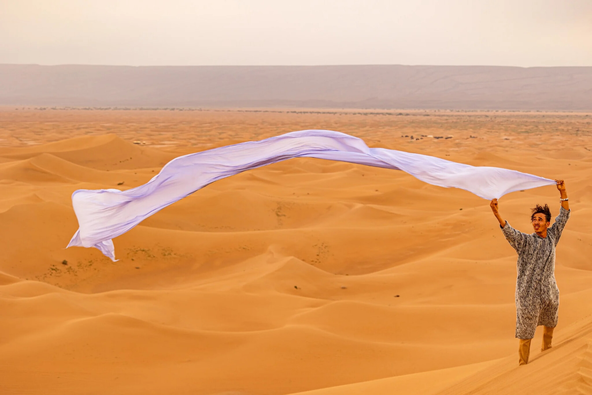 A young Moroccan host laughing as a long white scarf catches the wind above the dunes