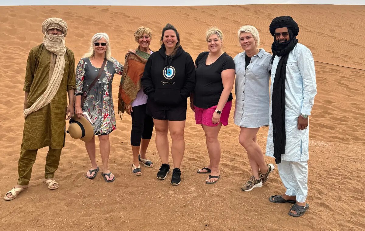 Hosts in desert robes standing with a group of smiling guests on the sand at Erg Chebbi