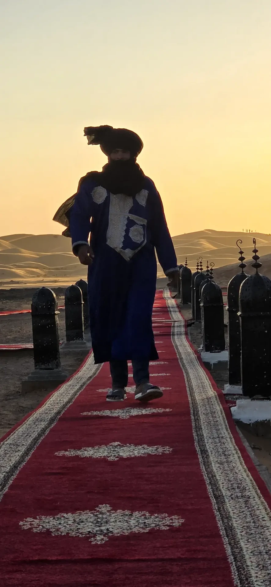 A host in a blue desert robe walking the lantern-lined carpet path as the sun sets over Erg Chebbi