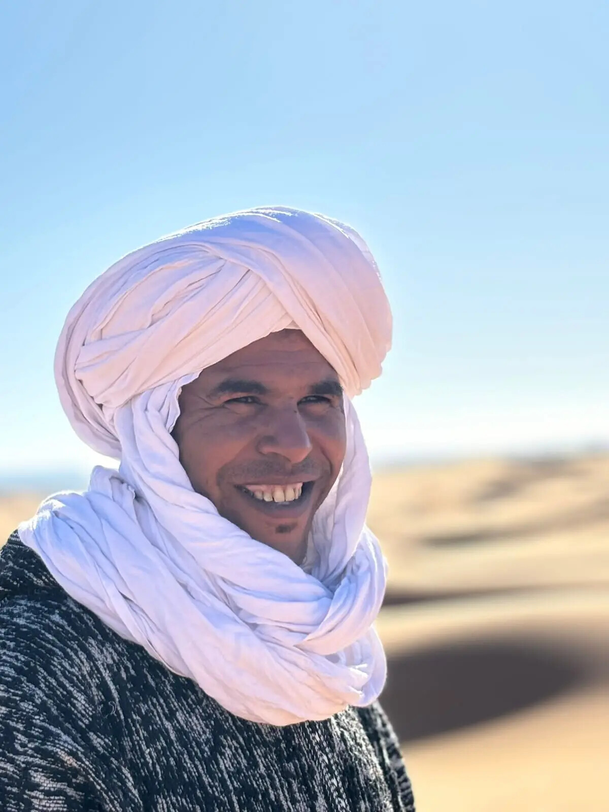 Ali, co-founder of Arabian Nights Desert Camp, smiling in a white turban with the dunes behind him