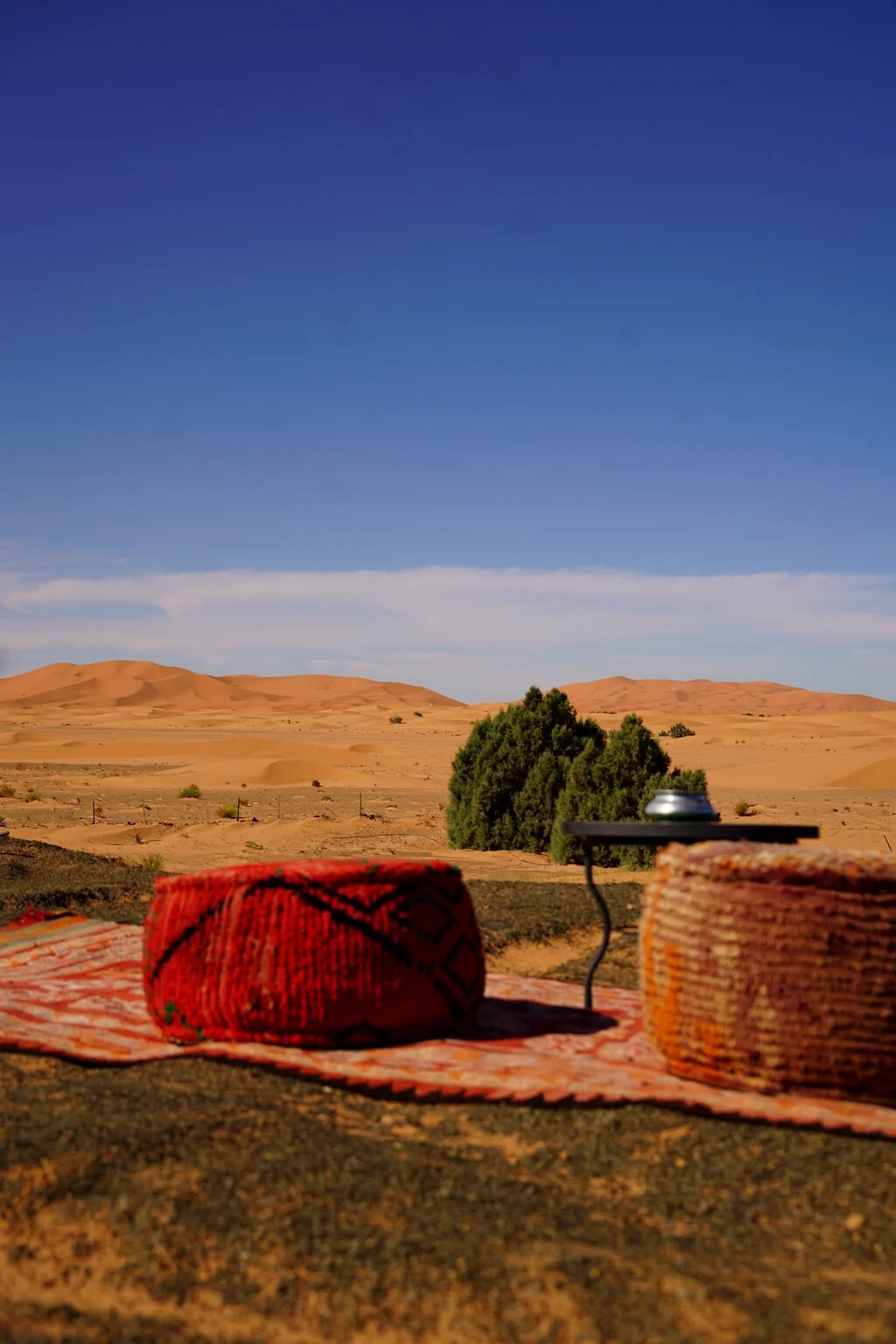 Woven poufs and a tea table on a rug facing the open Erg Chebbi dunes under a deep blue sky